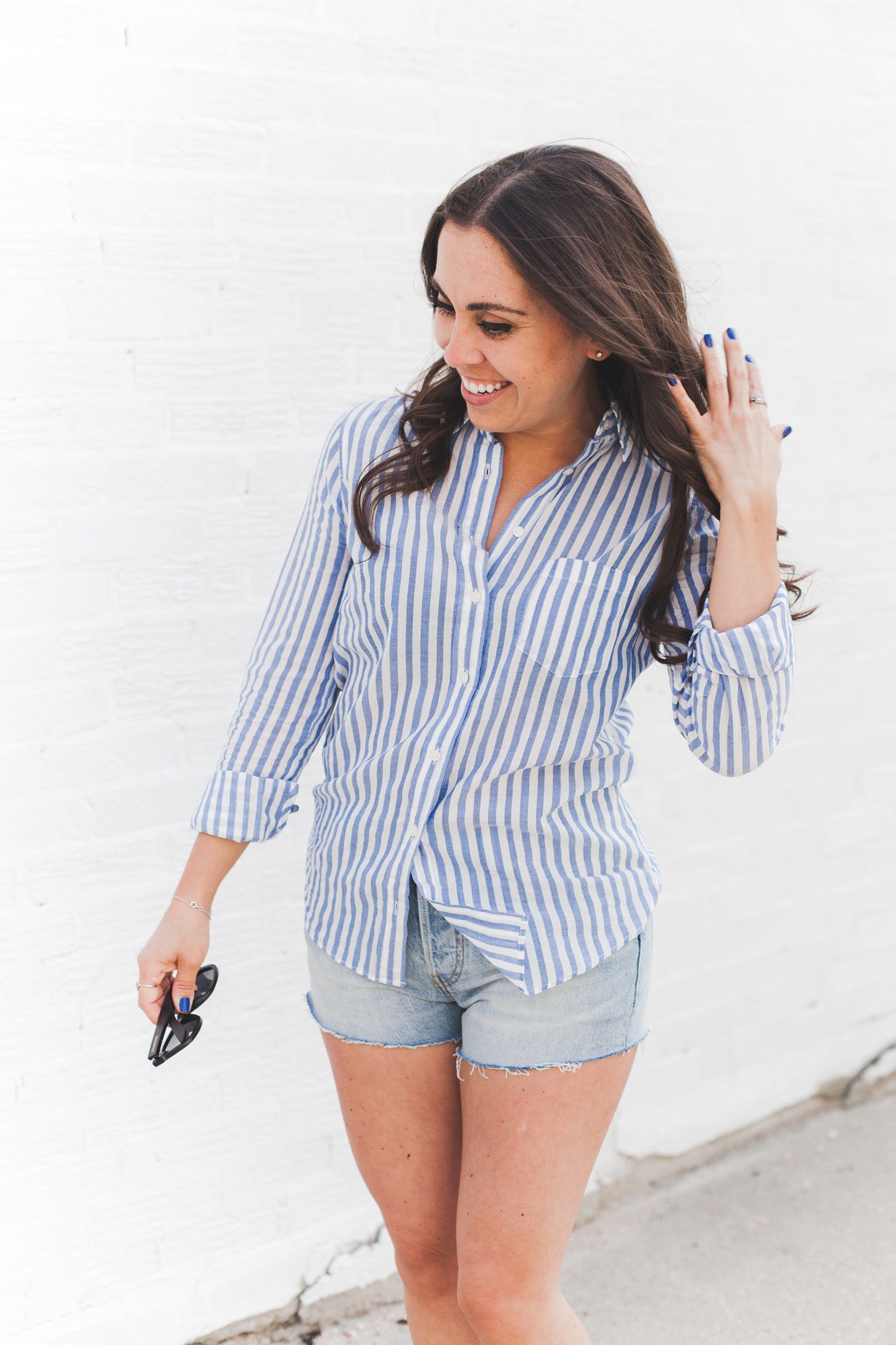 Smiling brunette woman in blue striped shirt and denim shorts walking near a white brick wall.