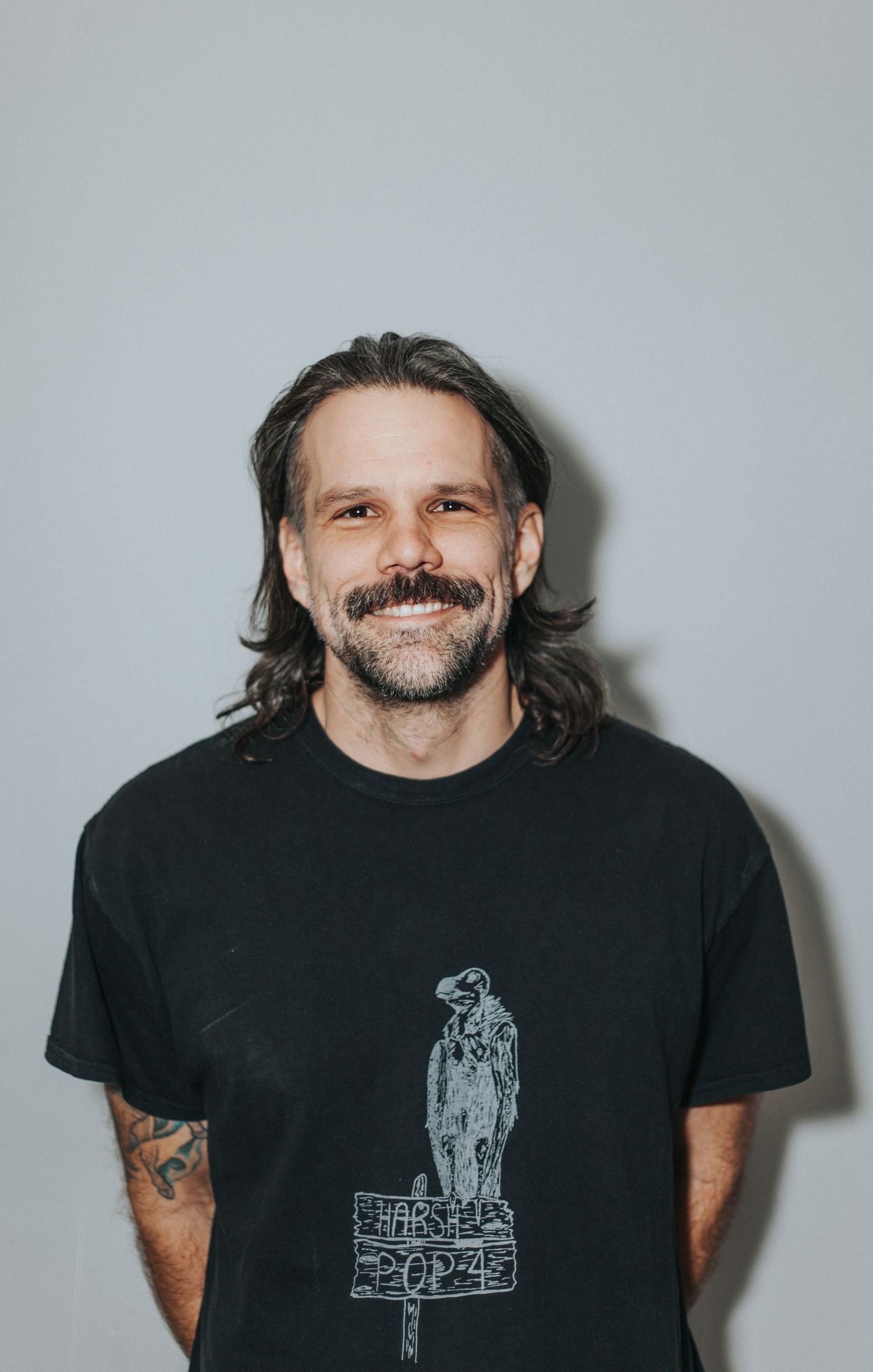 Smiling man with shoulder-length hair and mustache wearing a black graphic t-shirt.