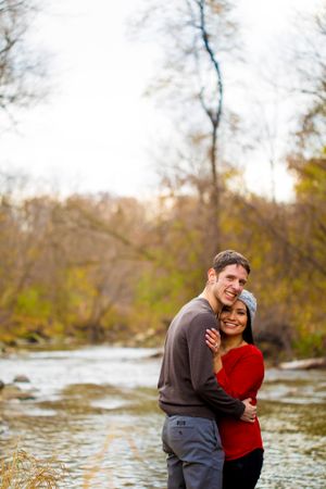 A couple embraces near a flowing river on an autumn day.