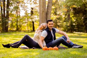 A couple relaxes on grass with orange pumpkins during an autumn photoshoot.