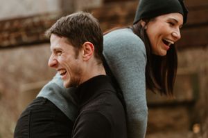 Playful couple laughing during piggyback ride outdoors in casual winter attire.