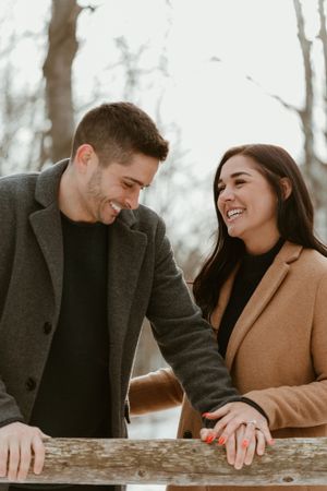 Couple in winter coats sharing genuine laughter while leaning on wooden railing.