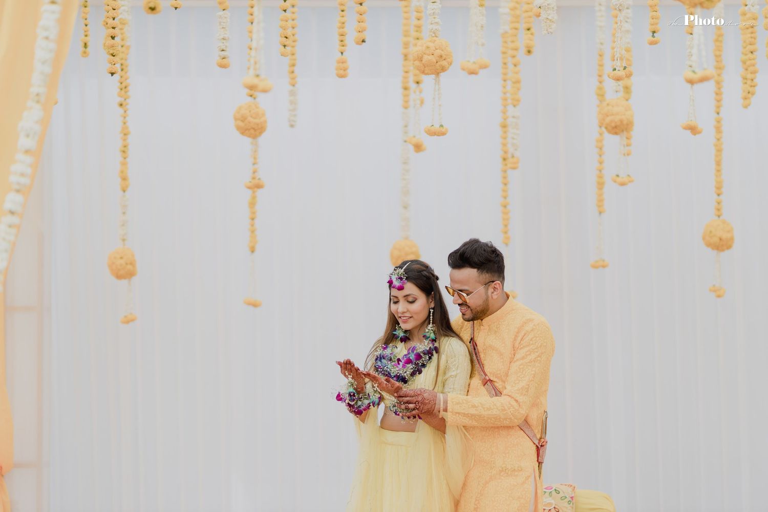 Couple in yellow traditional Indian attire performing rituals under hanging marigold decorations.