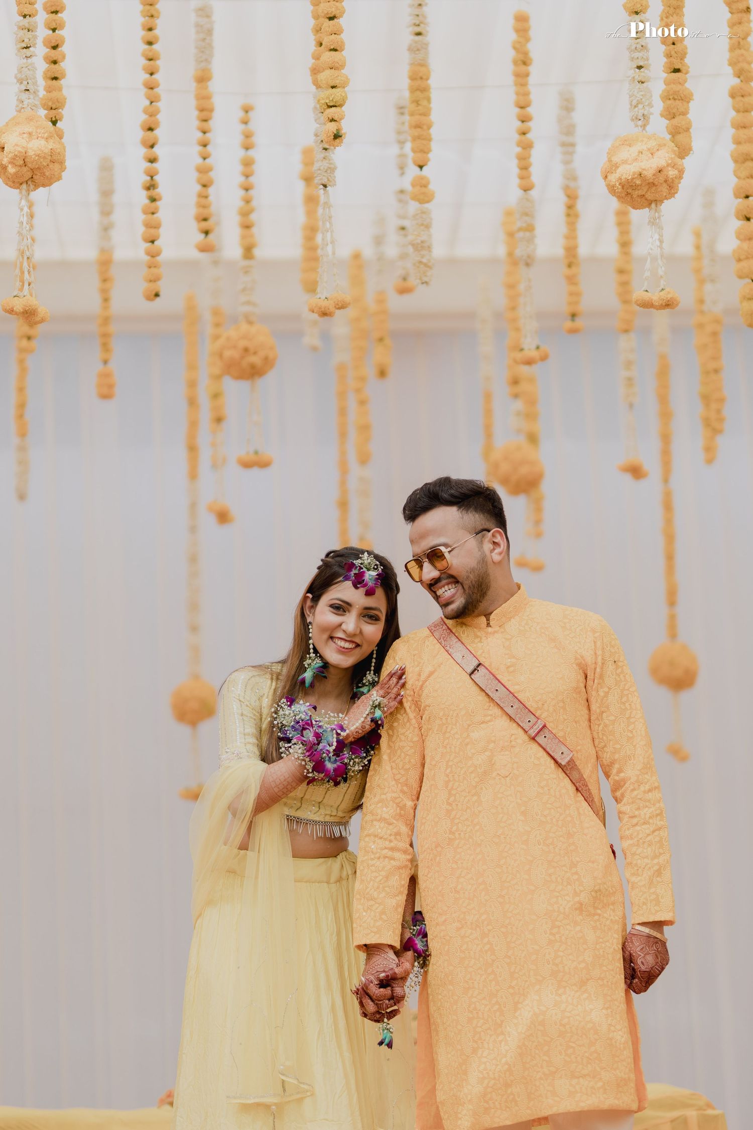 Couple in coordinated yellow outfits smiling under hanging floral decorations at Indian wedding ceremony.