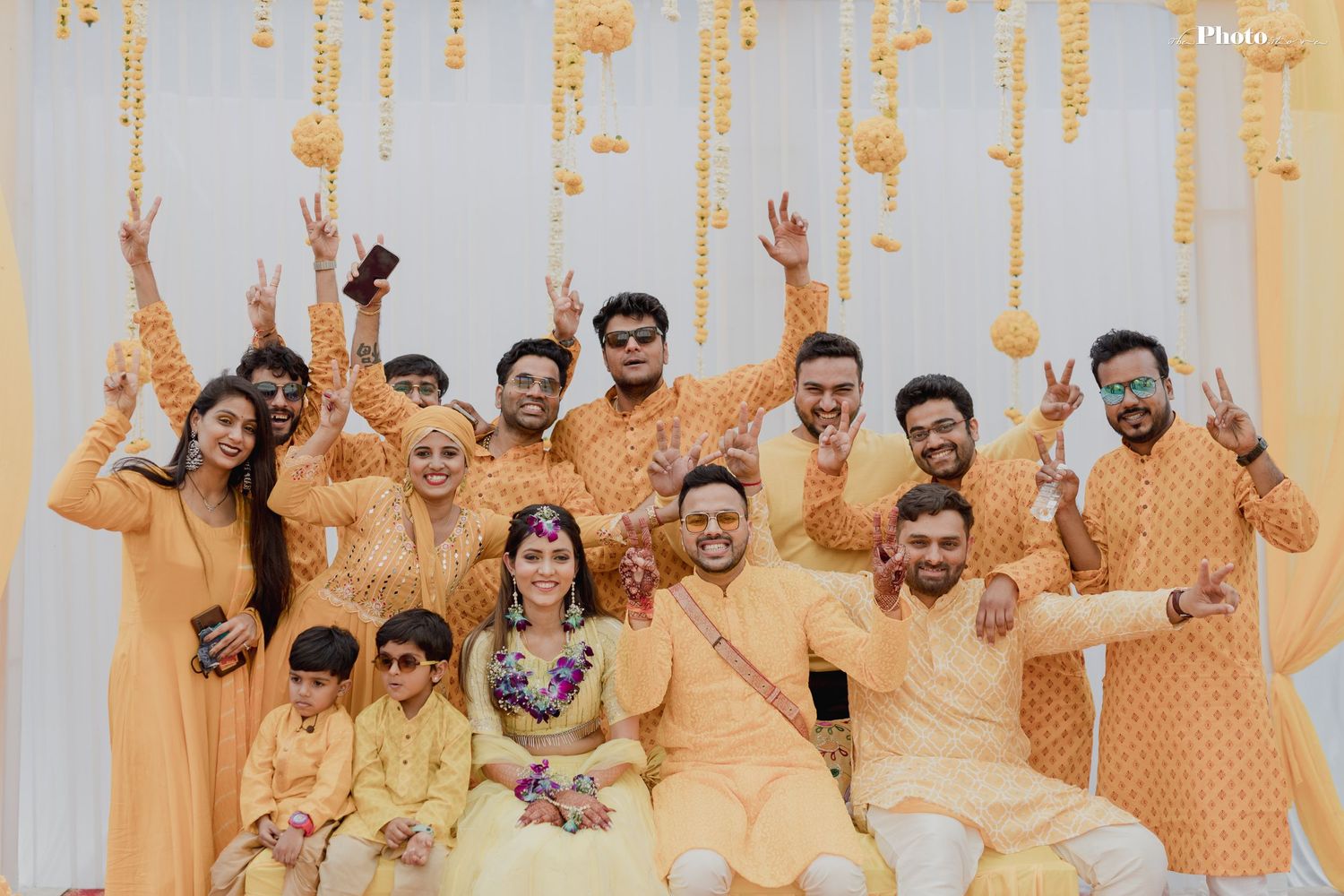 Large group in matching yellow outfits pose joyfully under hanging marigold decorations.