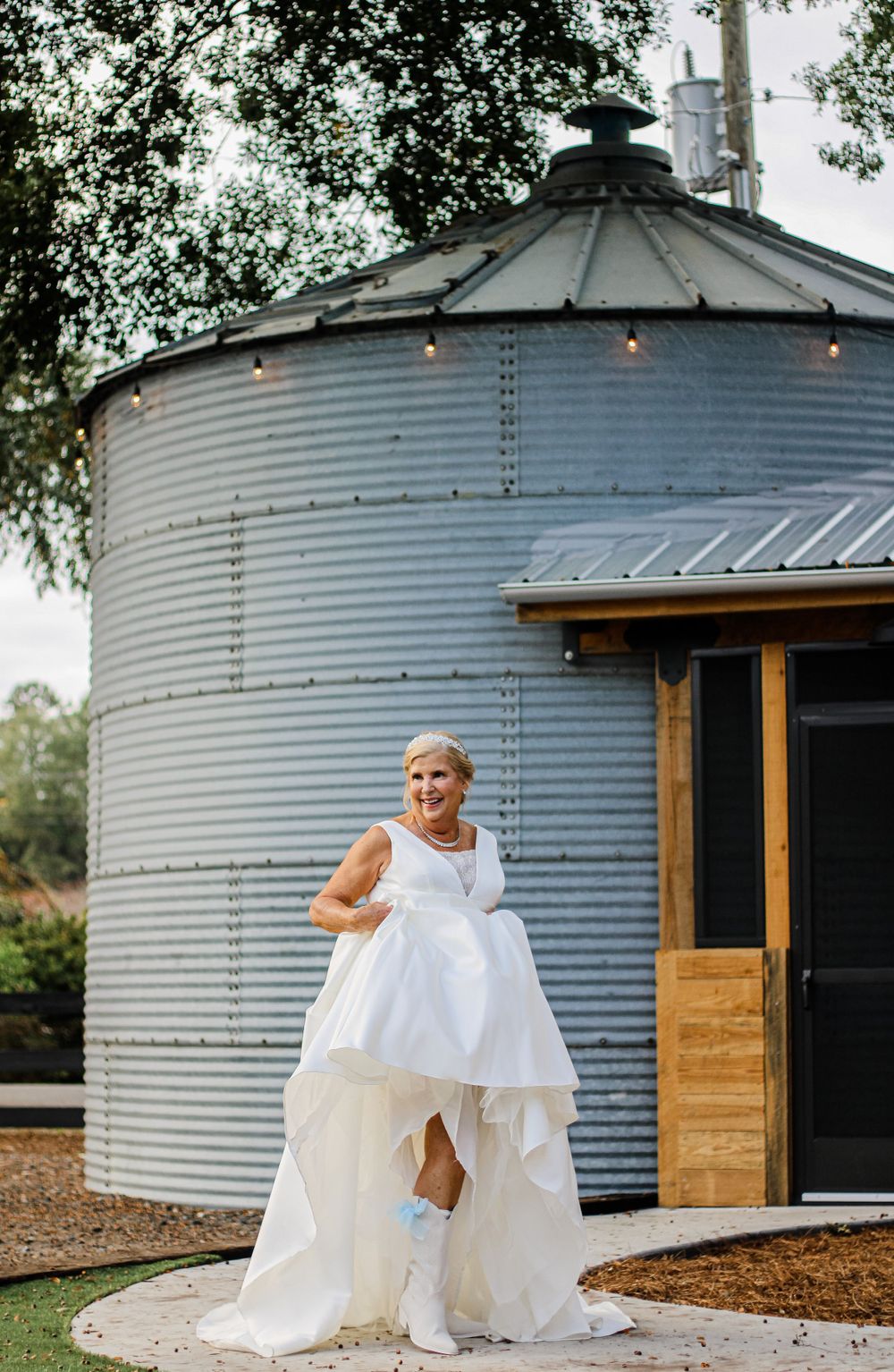Bride in white dress showing off boots while posing by rustic metal silo venue.