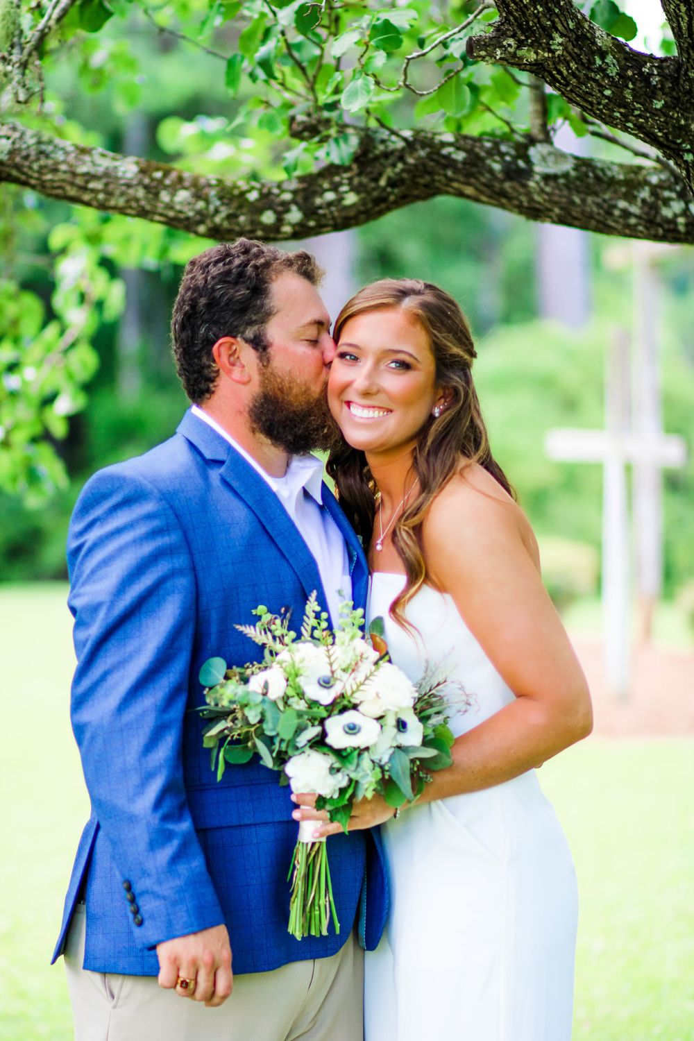 Romantic wedding couple embracing under tree with bride holding white floral bouquet.