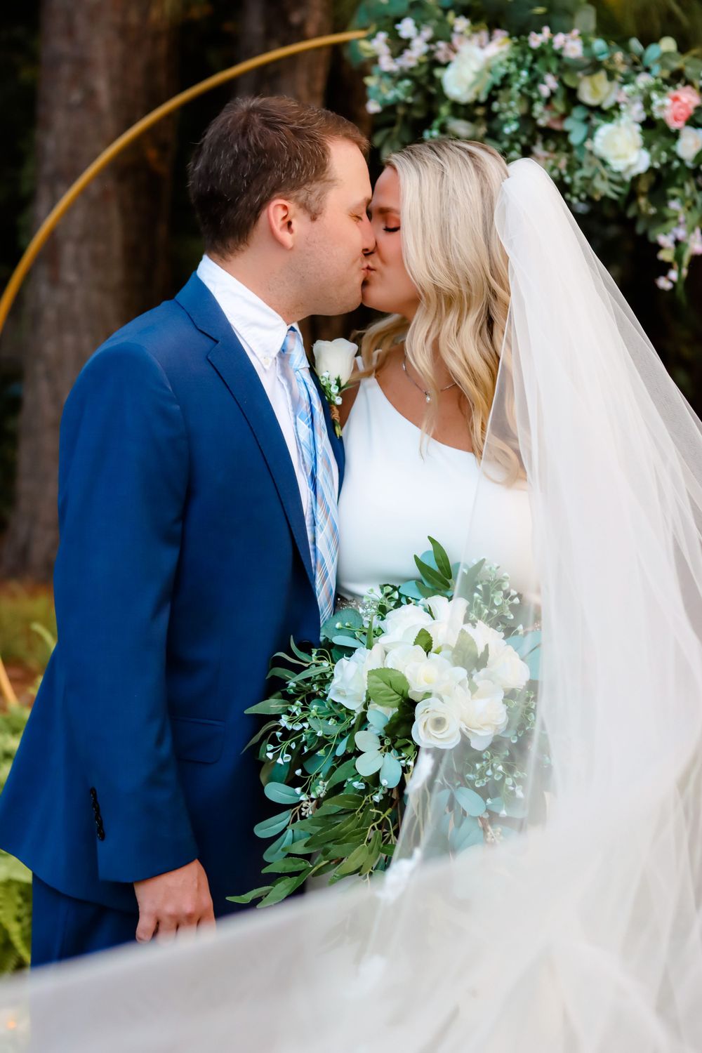 A romantic wedding kiss captured under a floral arch with white and green flowers.