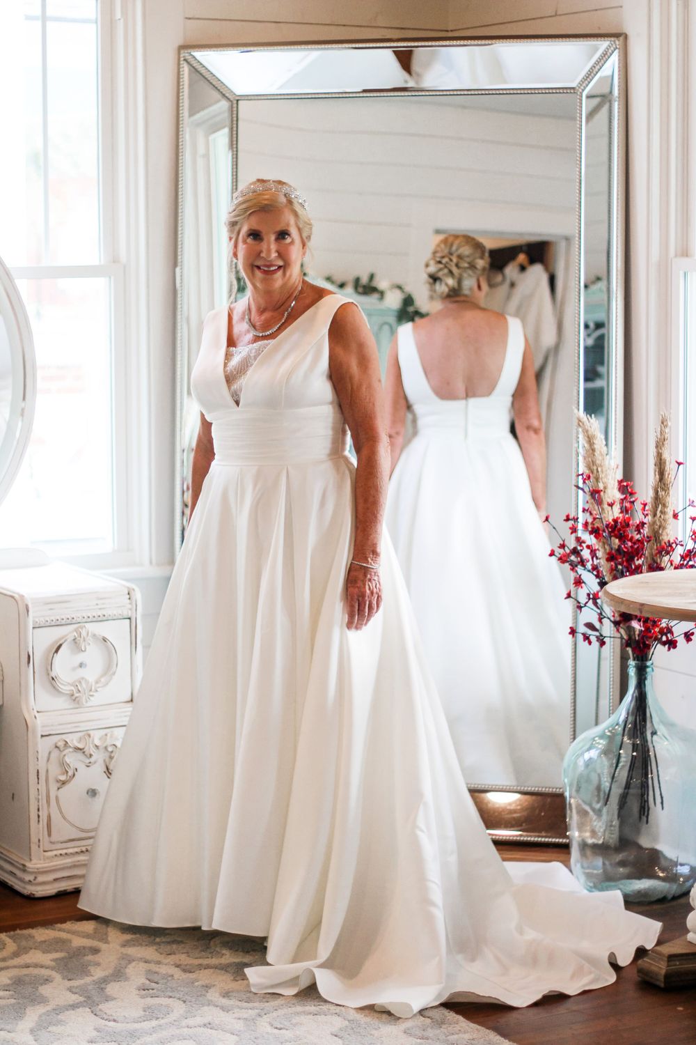 A bride in an elegant white dress stands beside a decorative mirror in a bright room.