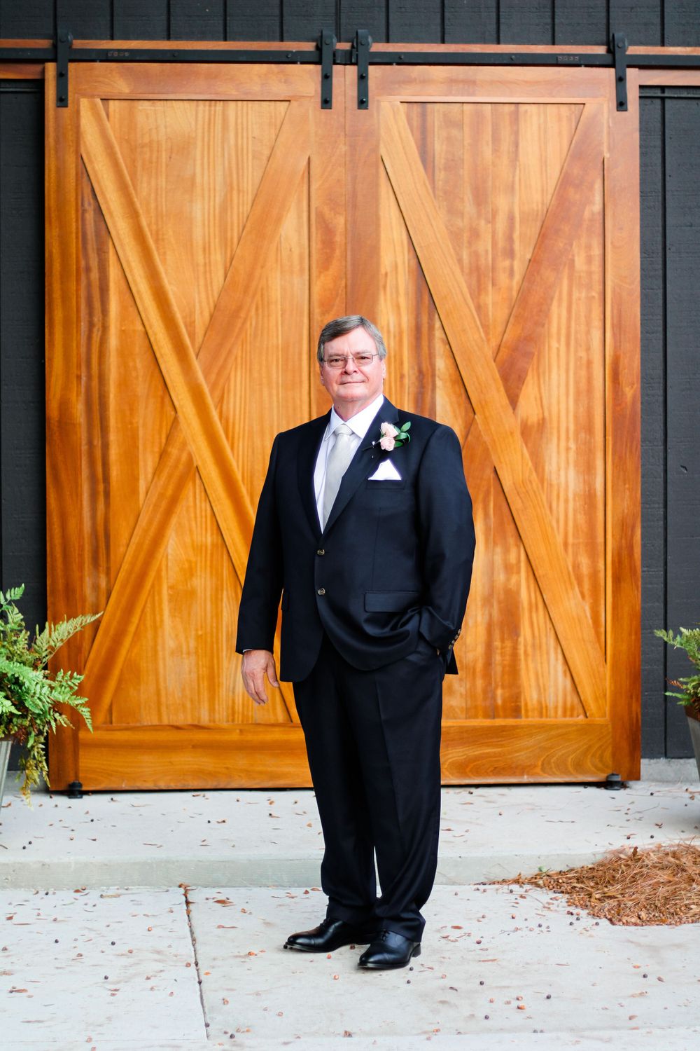 Groom in formal black tuxedo poses in front of wooden barn doors.