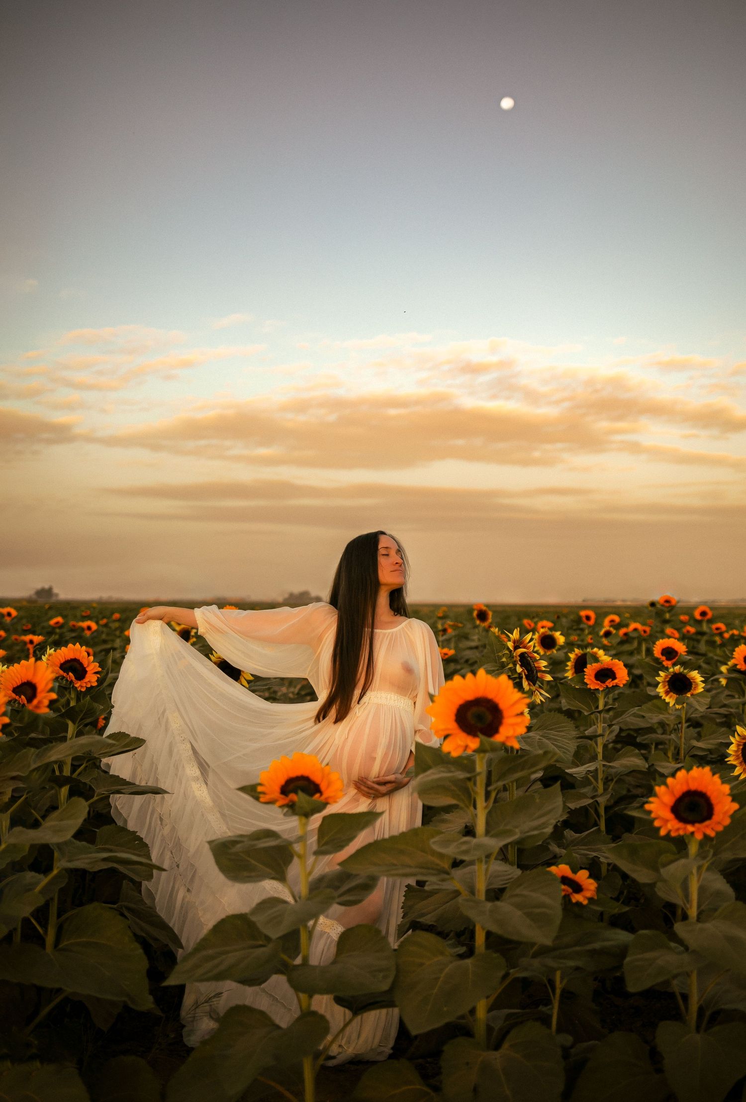 Figure in flowing white dress posing among sunflowers at sunset.