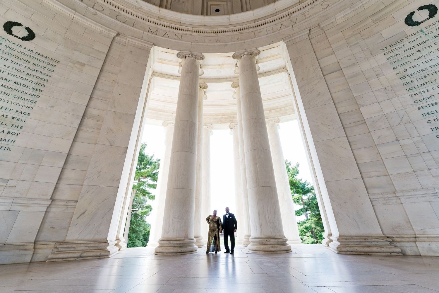 Silhouettes of two people stand within a grand marble rotunda with tall columns and natural light streaming in.