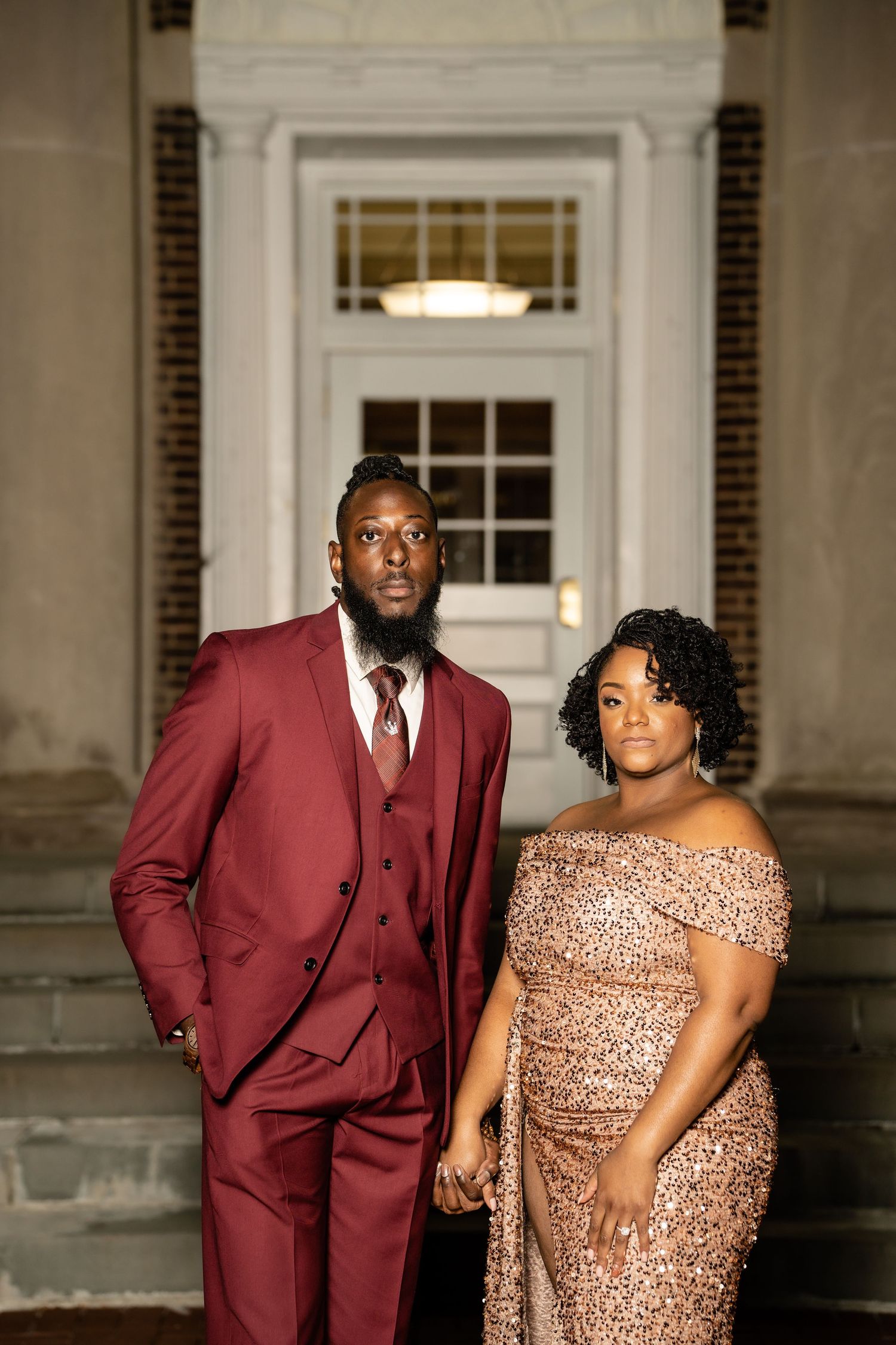 Elegant couple poses in formal attire with burgundy suit and sequined dress against classic architectural backdrop.