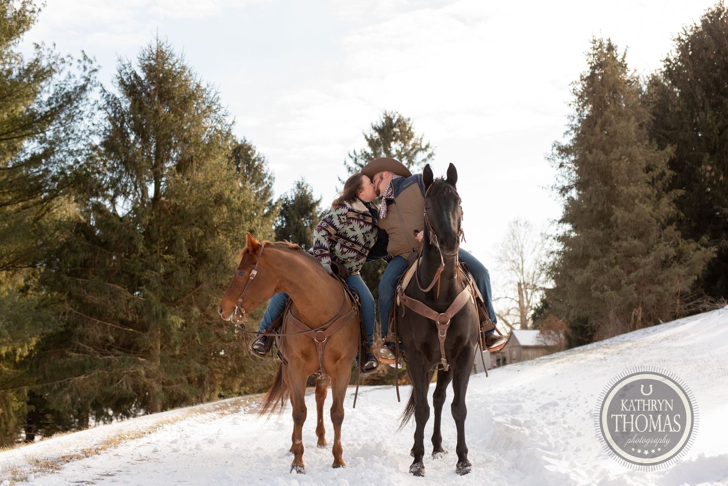 Two riders on horseback follow a snowy path through pine trees on a winter afternoon.