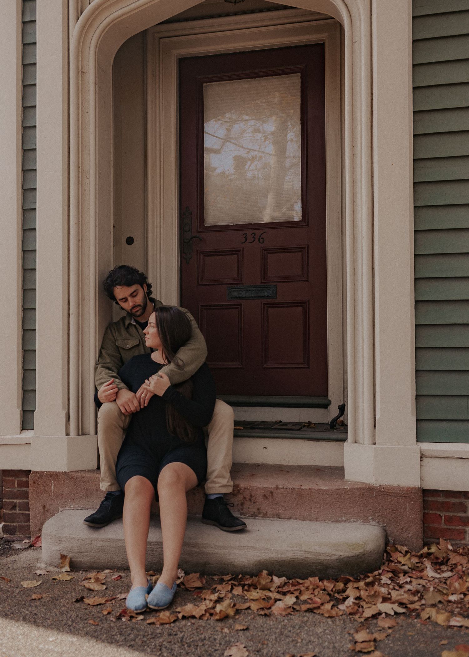 A couple sits together on concrete steps in front of a brown wooden door on an autumn day on Benefit St in Providence, RI.