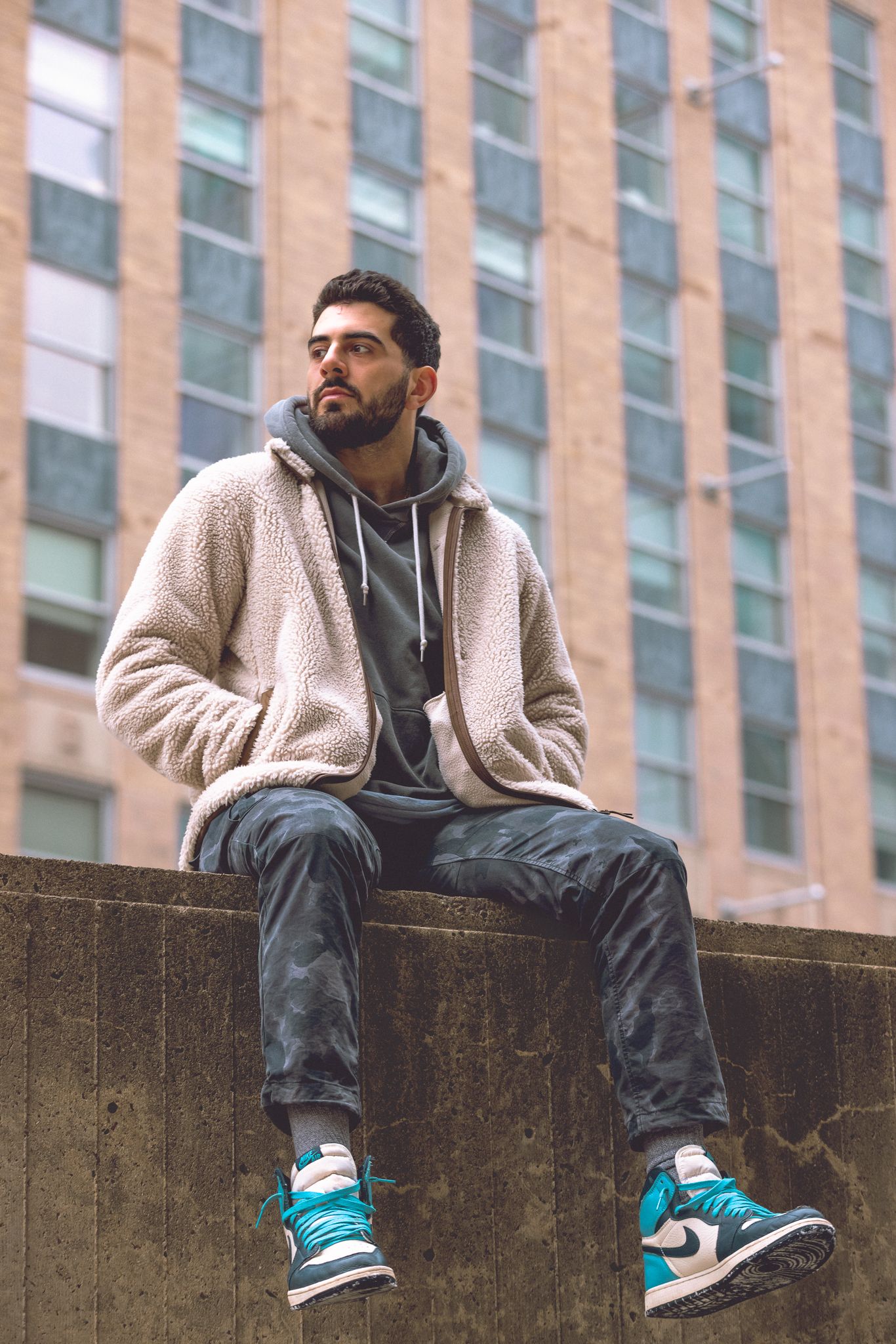 Person in casual streetwear sitting on concrete ledge against brick building backdrop.
