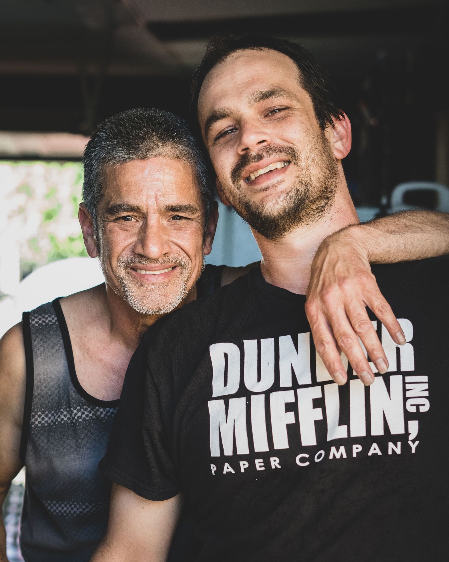 Two smiling men posing together, one wearing a Dunder Mifflin Paper Company t-shirt, in a casual outdoor setting.
