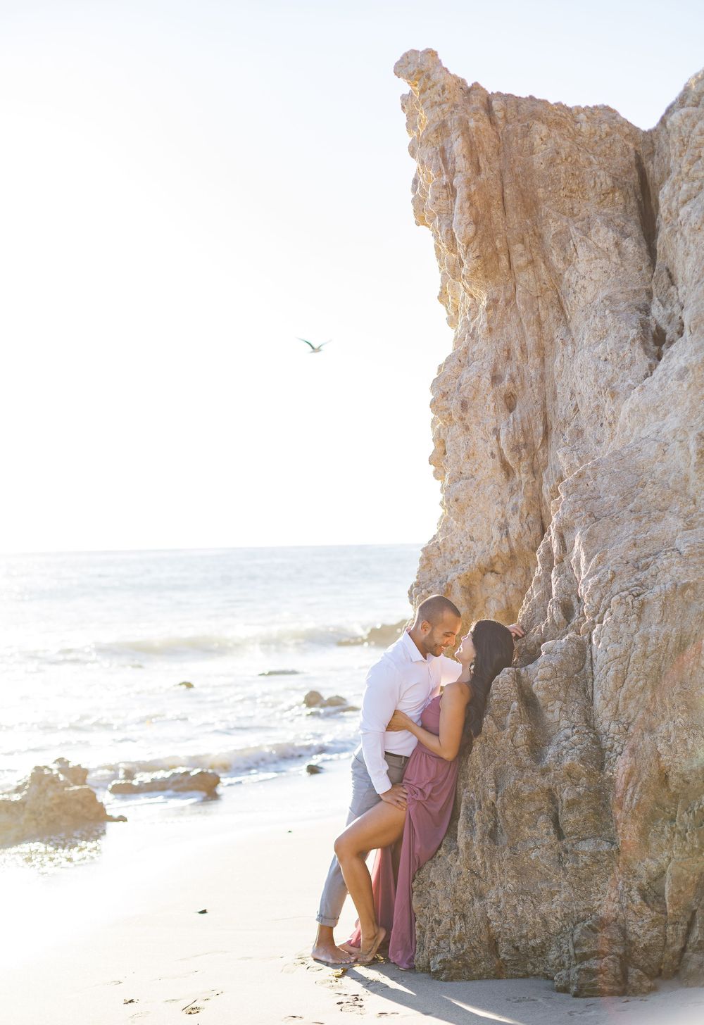 Intimate moment between couple against sunlit beach cliffs with ocean waves in background.