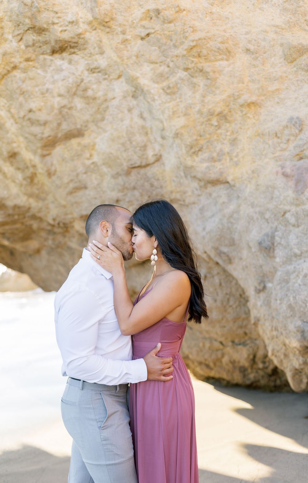 A loving couple shares an intimate moment on the beach beside rocky cliffs.