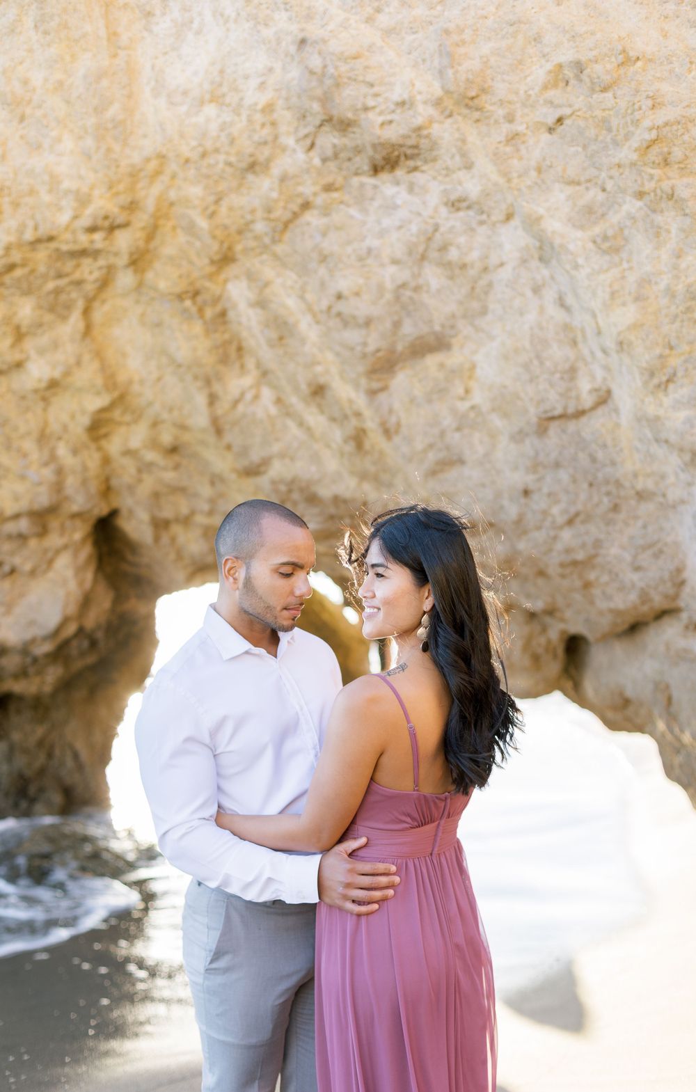 Romantic couple in pink dress and white shirt embrace by coastal rock formations.