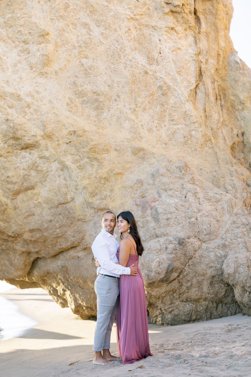 Couple poses together against towering beige coastal cliffs at the beach.
