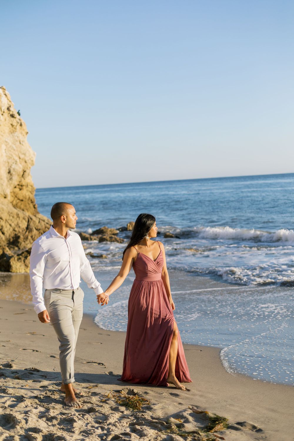 A couple strolls along the beach holding hands beside tall coastal cliffs.