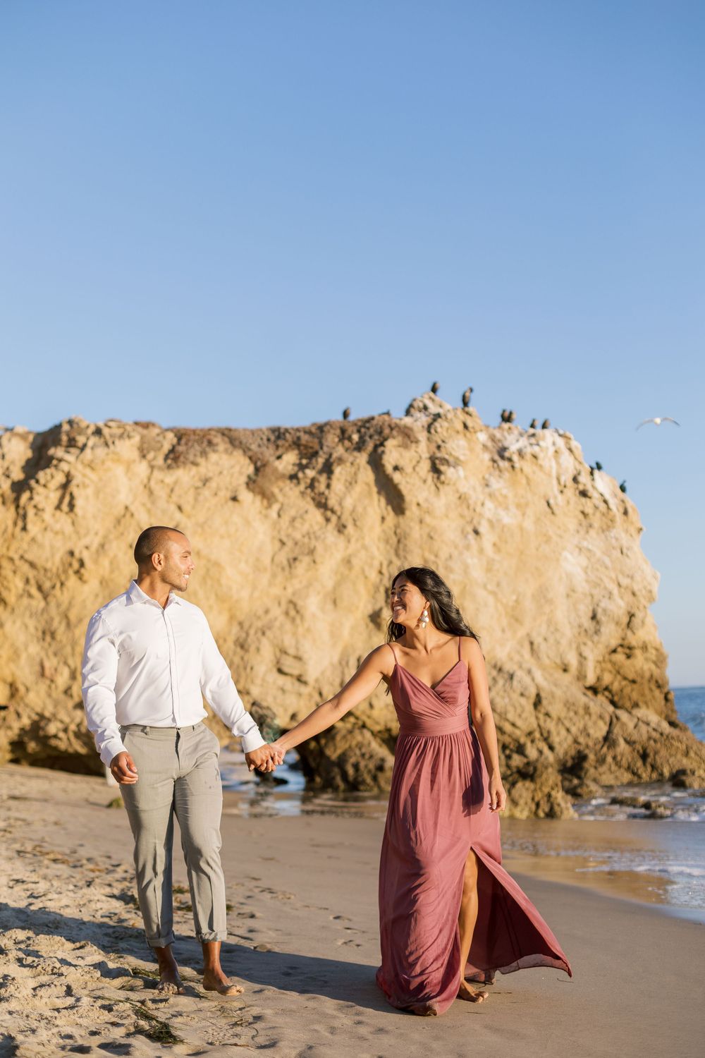 A couple walks hand in hand on a sandy beach with dramatic cliffs and seabirds in the background.