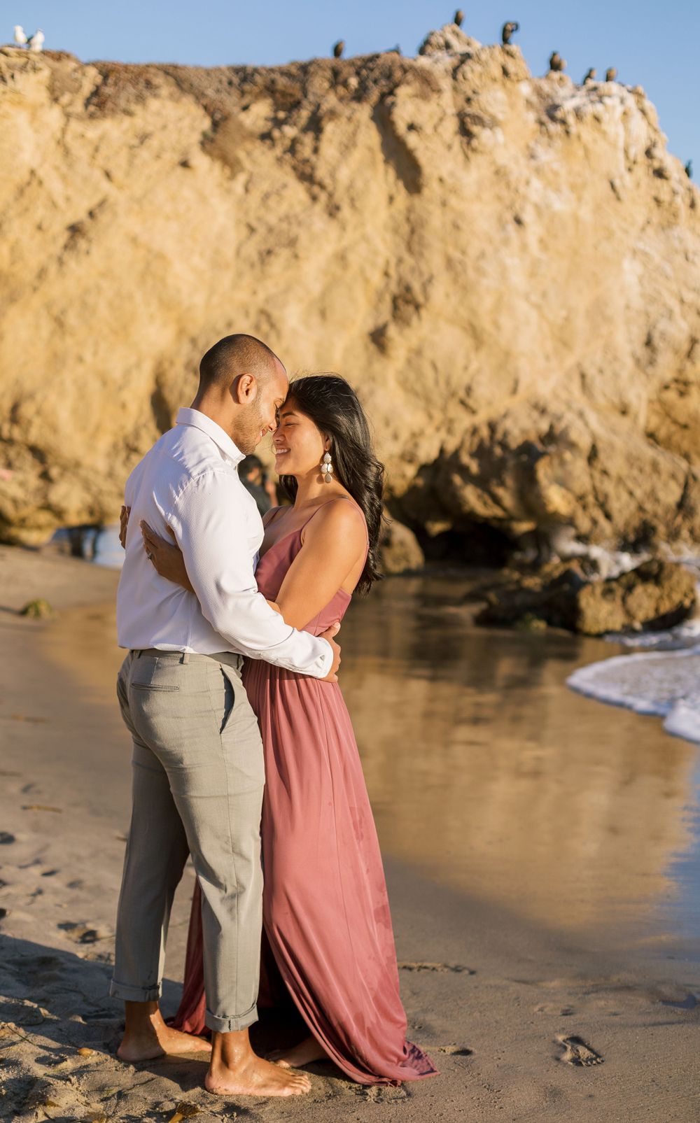 A couple shares an intimate moment on the beach during golden hour with cliffs in the background.