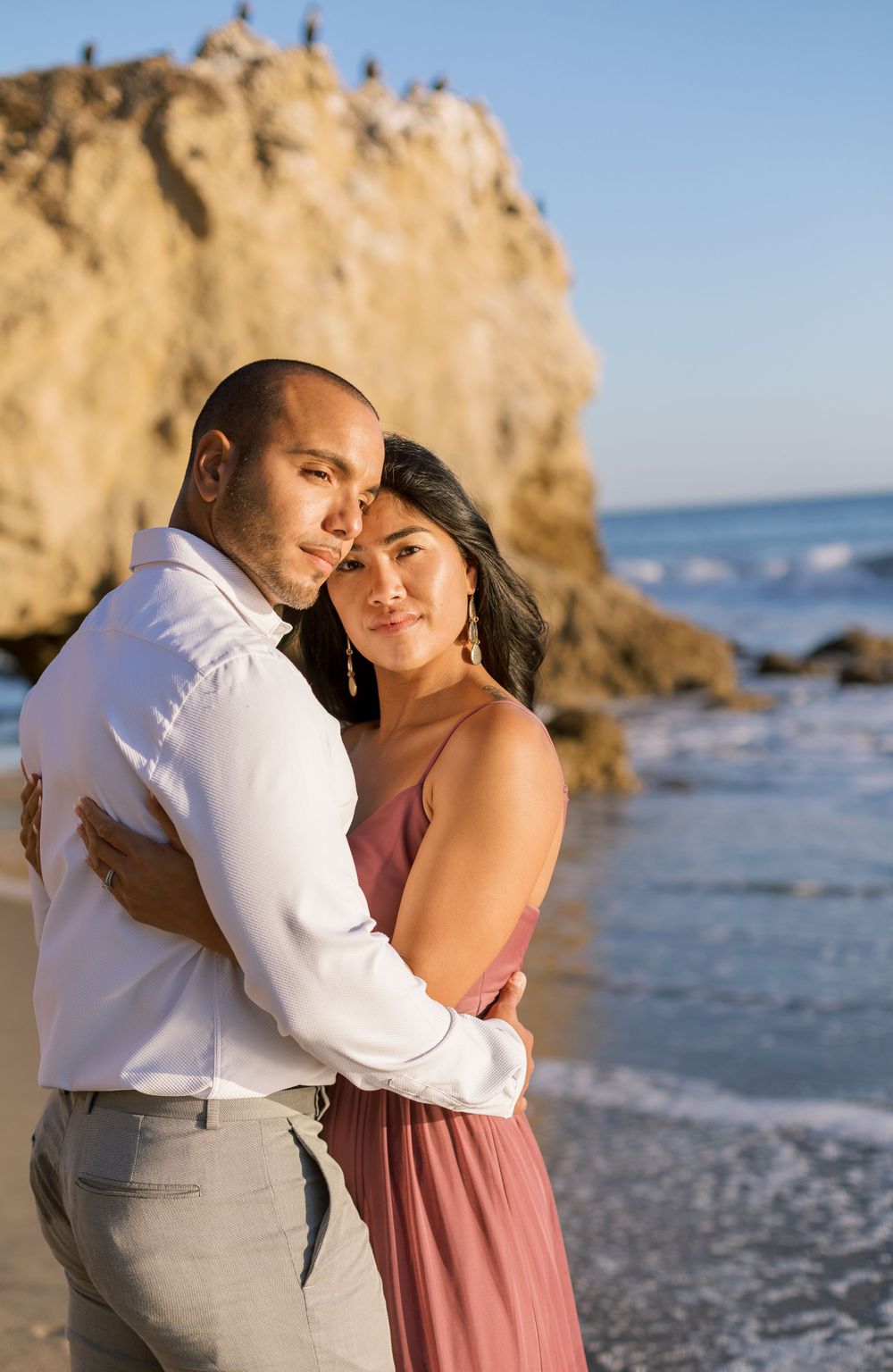 A romantic couple embraces on a beach near large coastal rocks during golden hour.