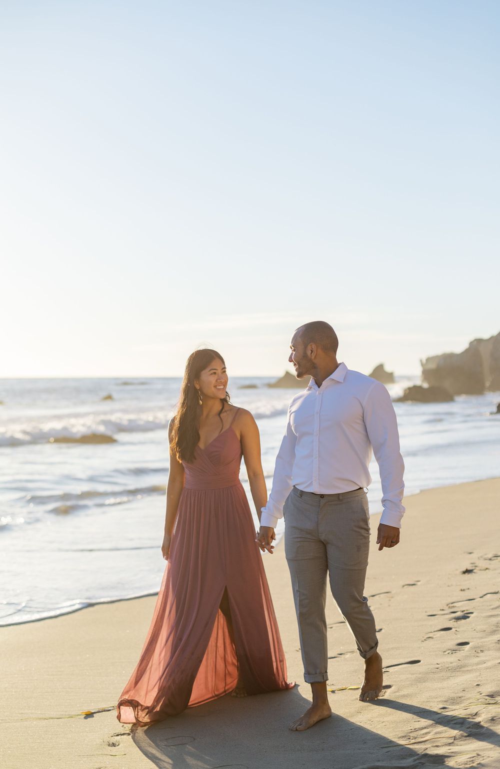 Couple walks along beach shoreline at sunset wearing formal attire.