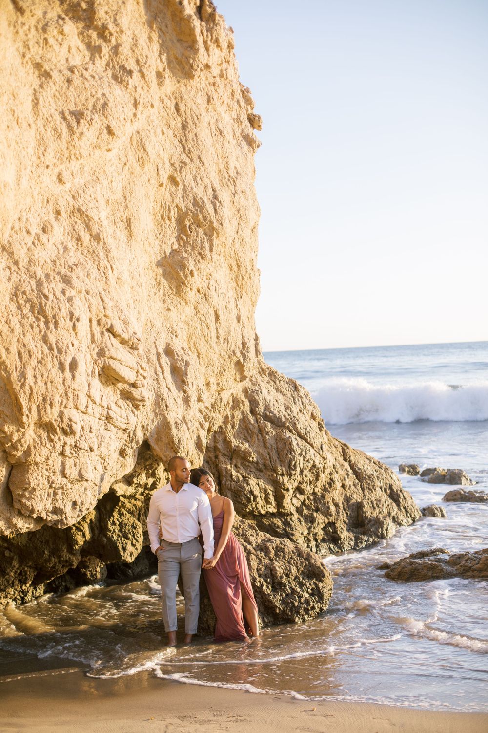 A couple stands together by a rocky cliff on a beach during sunset with waves crashing in the background.