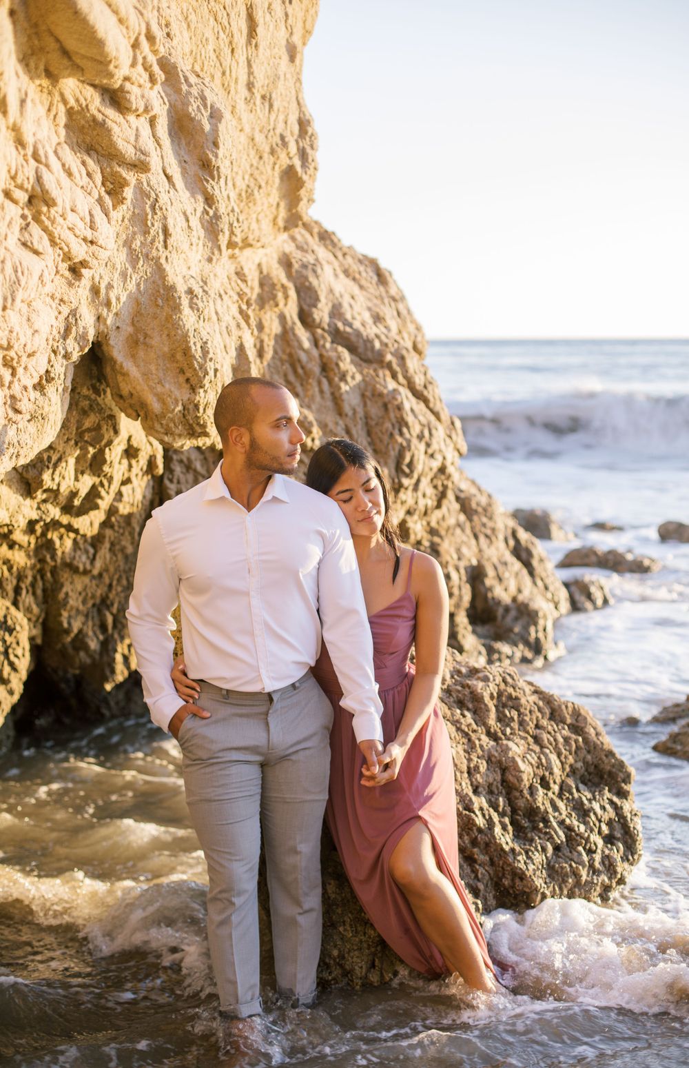 Couple poses against coastal rock formation as waves crash behind them.