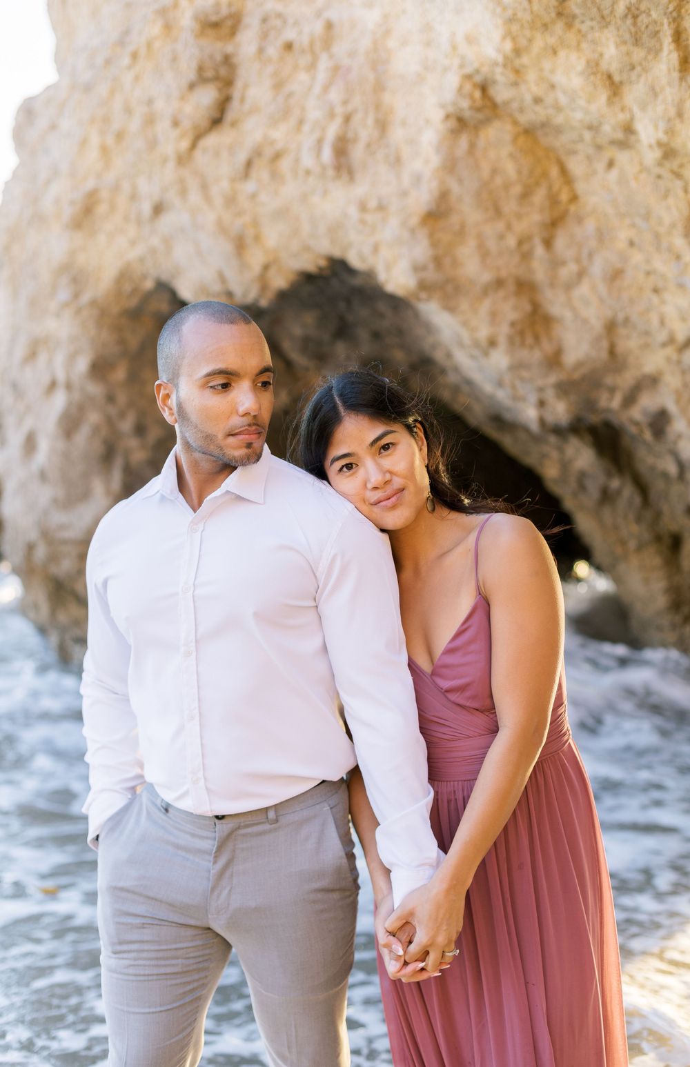 A couple in formal attire embracing by coastal rock formations at the beach.
