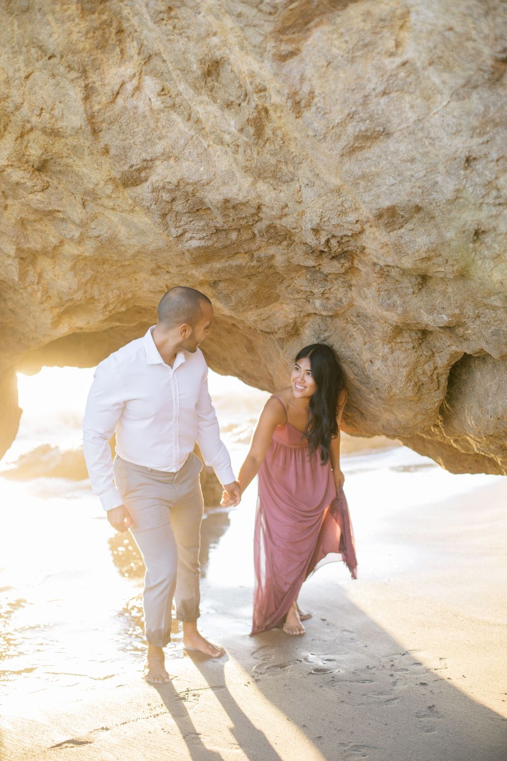 Couple walks hand in hand through beach cave with sunlight streaming in.