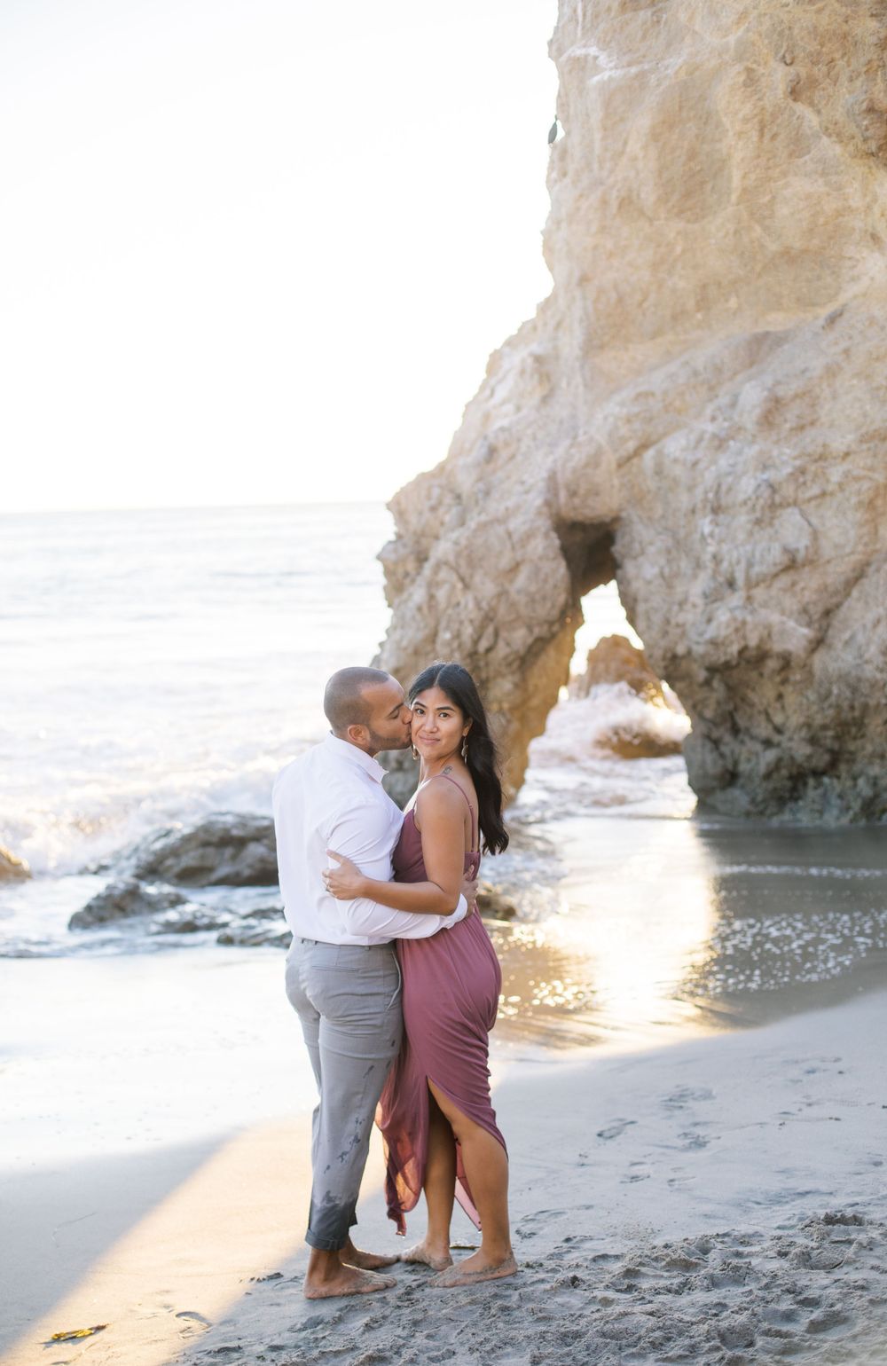 A sunset beach portrait of a couple embracing near a natural rock arch.