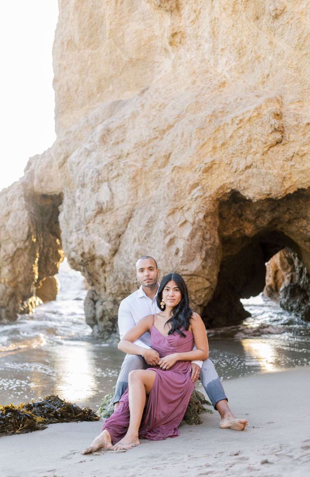 Romantic beach photoshoot with couple sitting on sand near natural rock arch.