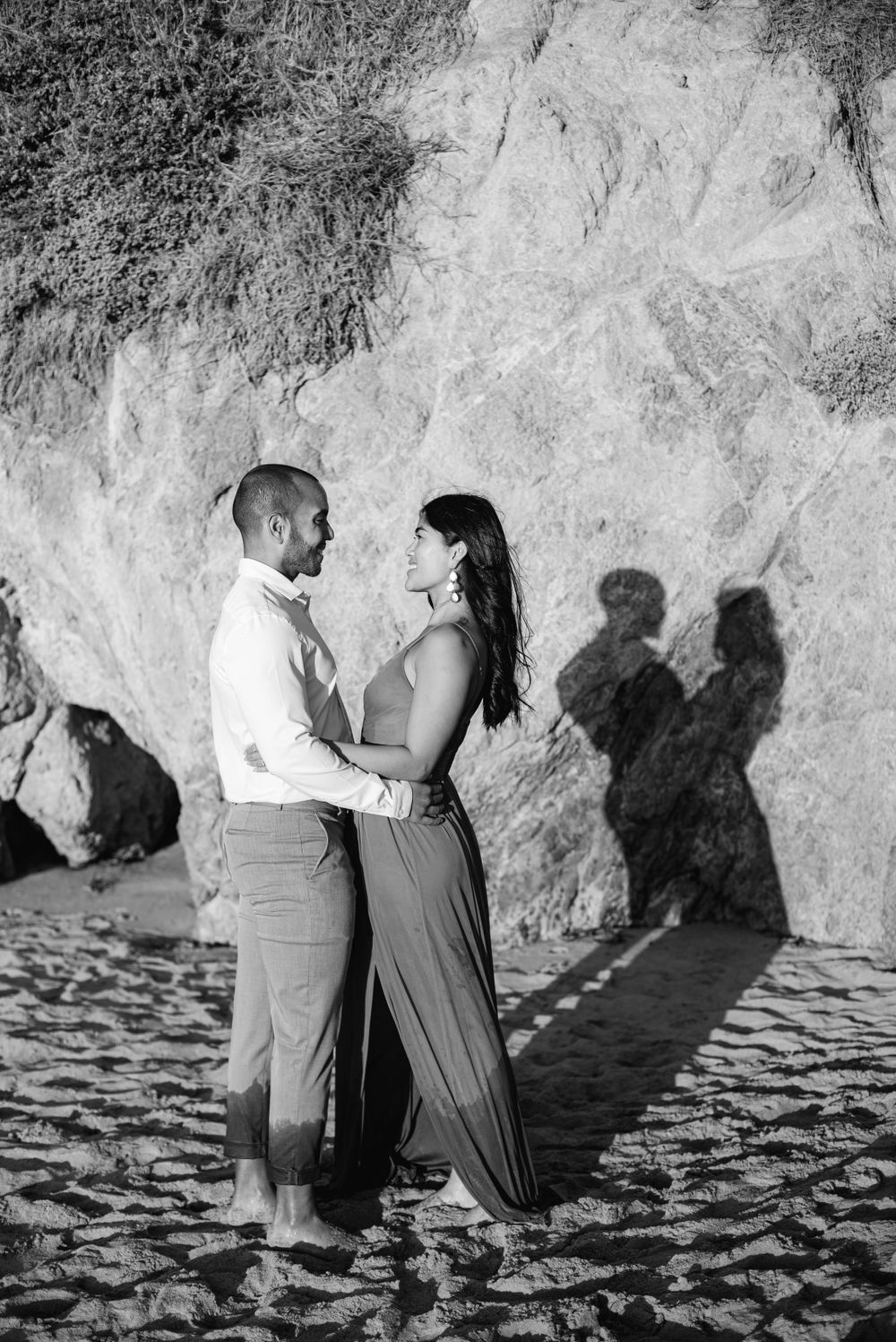 A couple creates shadows on the sand as they embrace near a cliff wall in black and white.