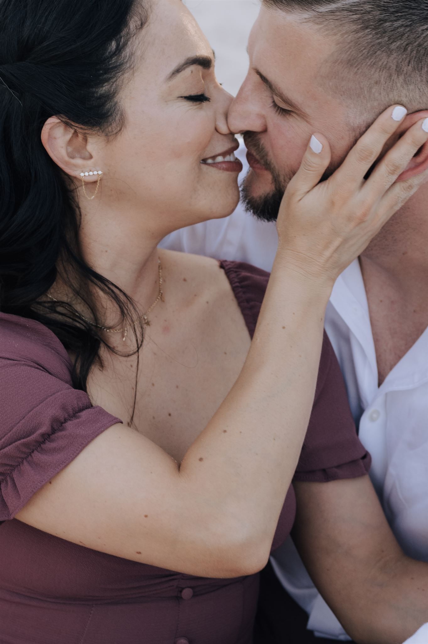 Close up of intimate moment between couple in burgundy dress and white shirt against natural backdrop.