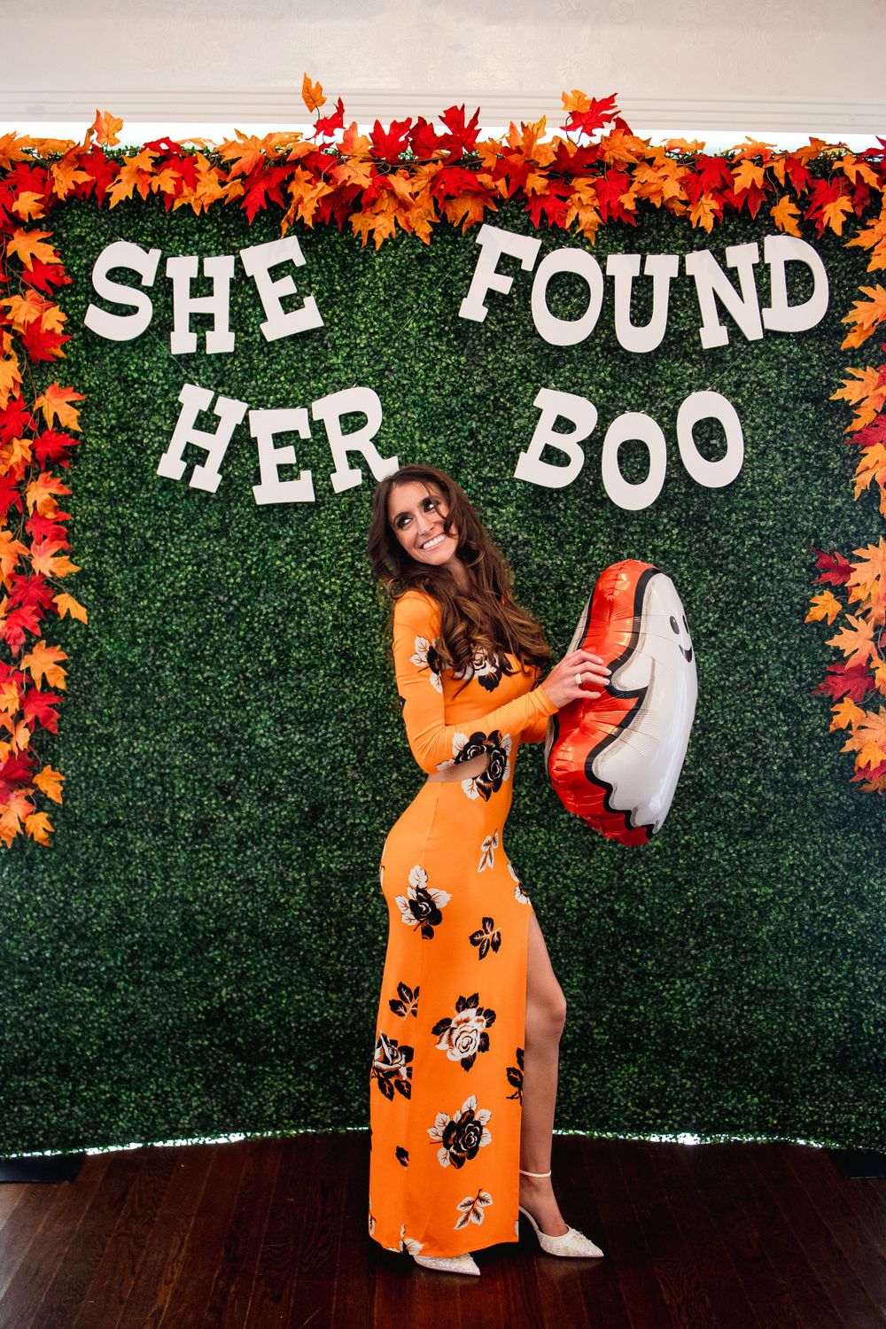 Woman in orange floral dress posing against decorative fall-themed photo backdrop.