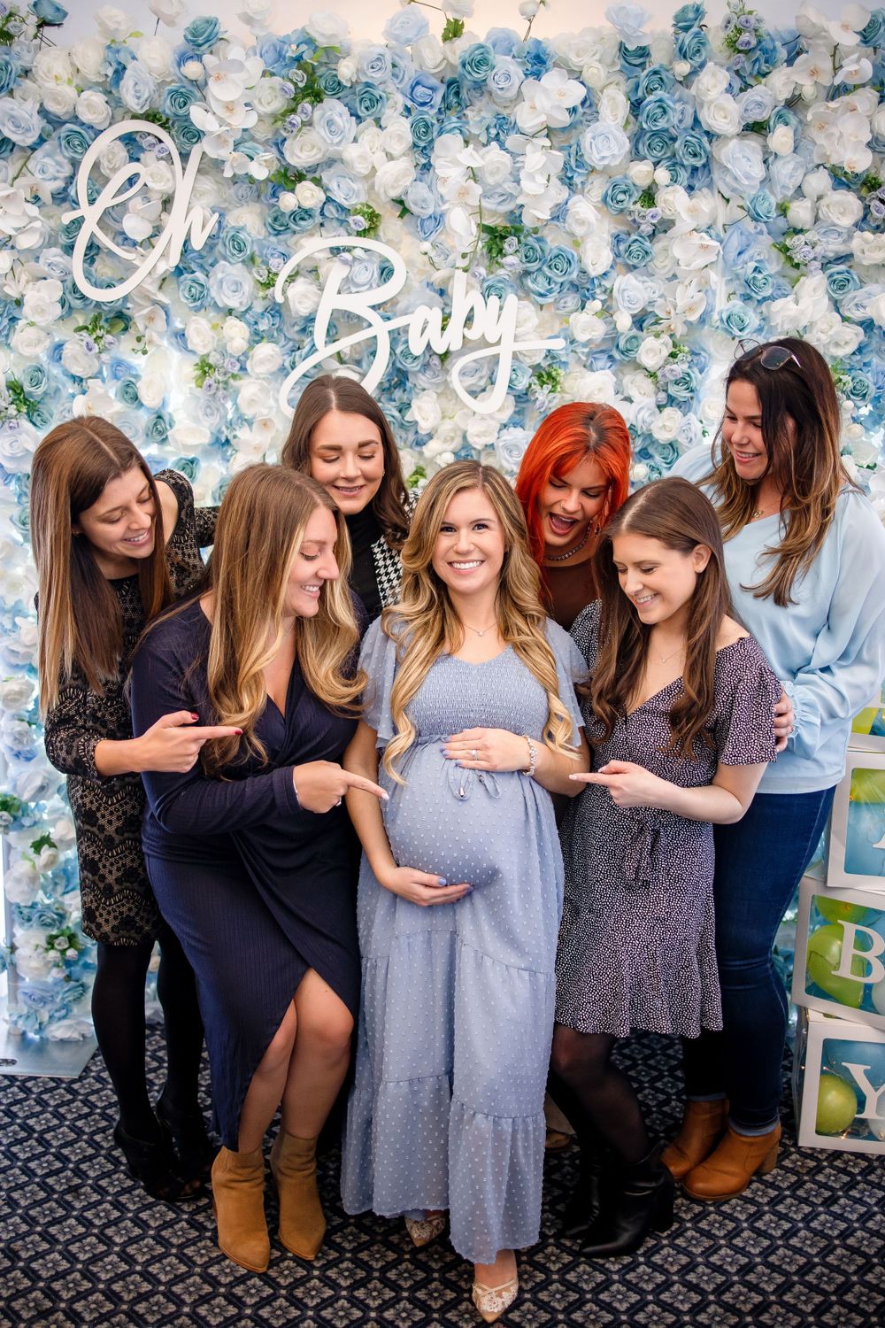 Group of friends gather around expectant mother at baby shower with floral backdrop.