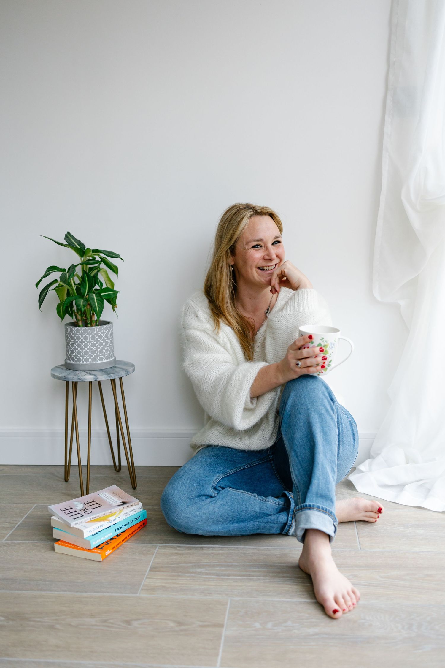 Woman in cozy sweater relaxes with coffee by window near houseplant and stack of books.