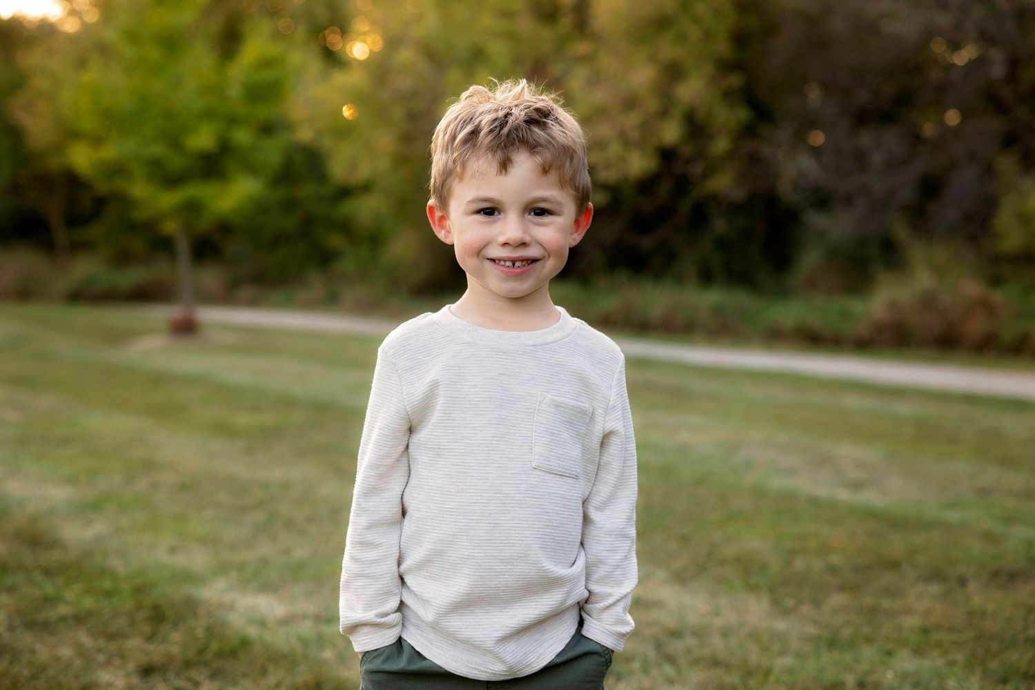Child wearing light colored sweater stands smiling in grassy outdoor setting at golden hour.