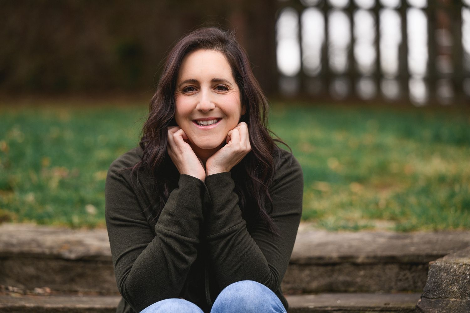Smiling brunette woman sitting on stone steps with hands on cheeks.
