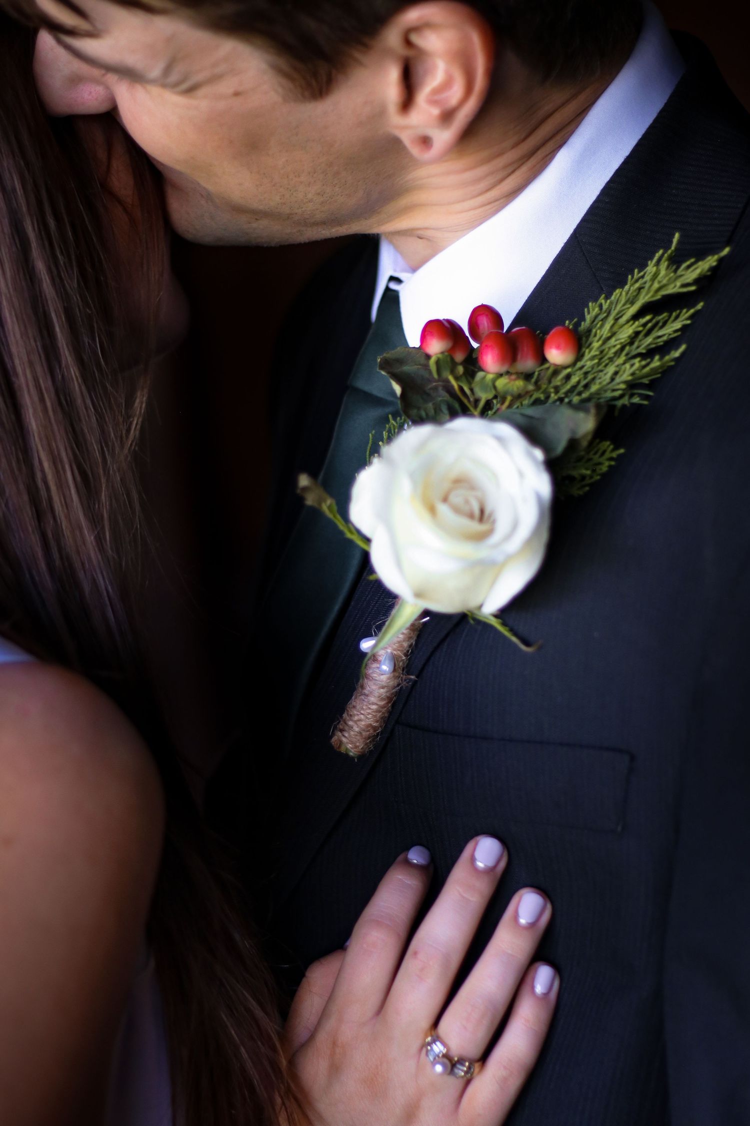Passionate couple at wedding, groom wearing black suit with white rose and red berry boutonniere, bride showing diamond ring.