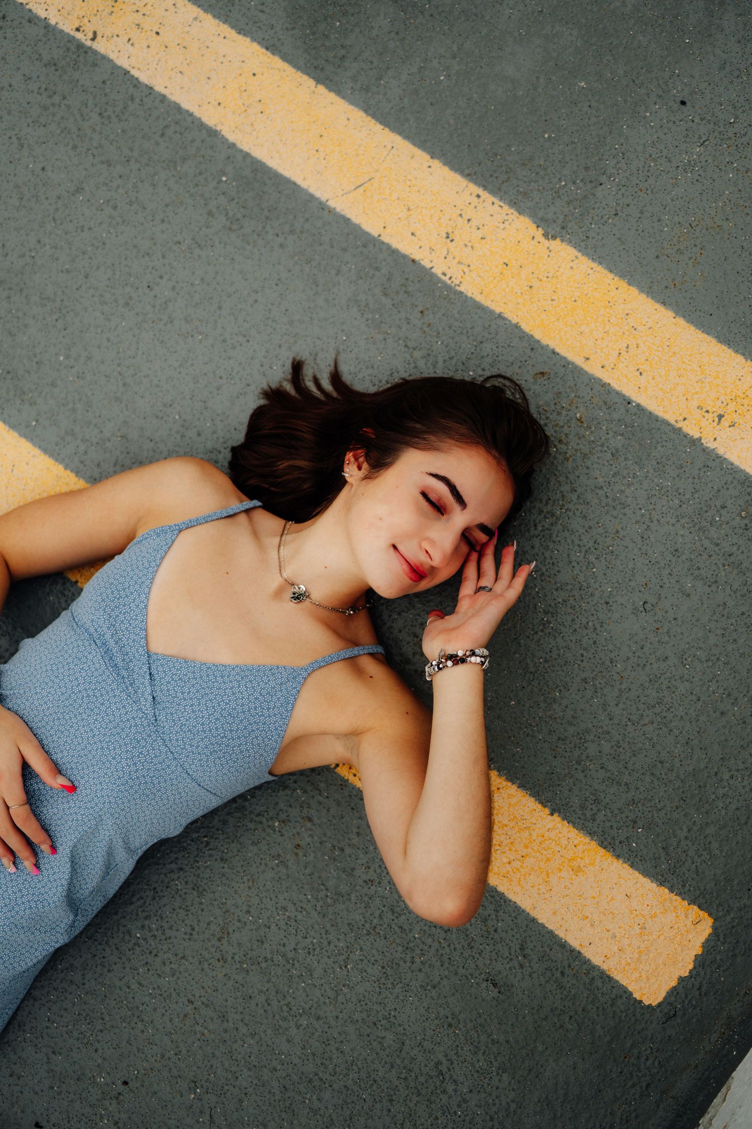Artistic overhead shot of someone lying on pavement with yellow line, wearing blue dress and dark hair.