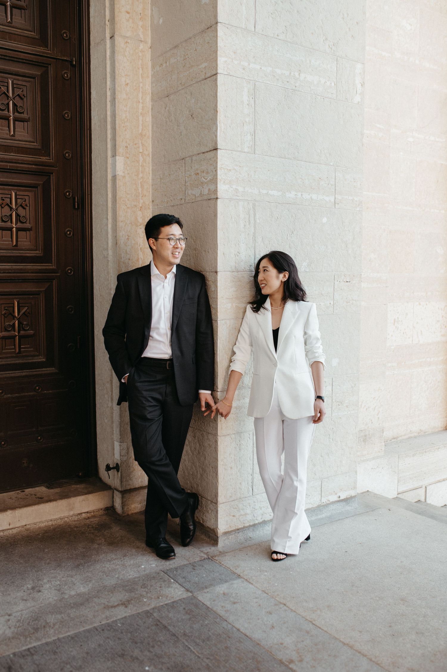 Elegant couple smiling and holding hands while walking by a grand stone wall.