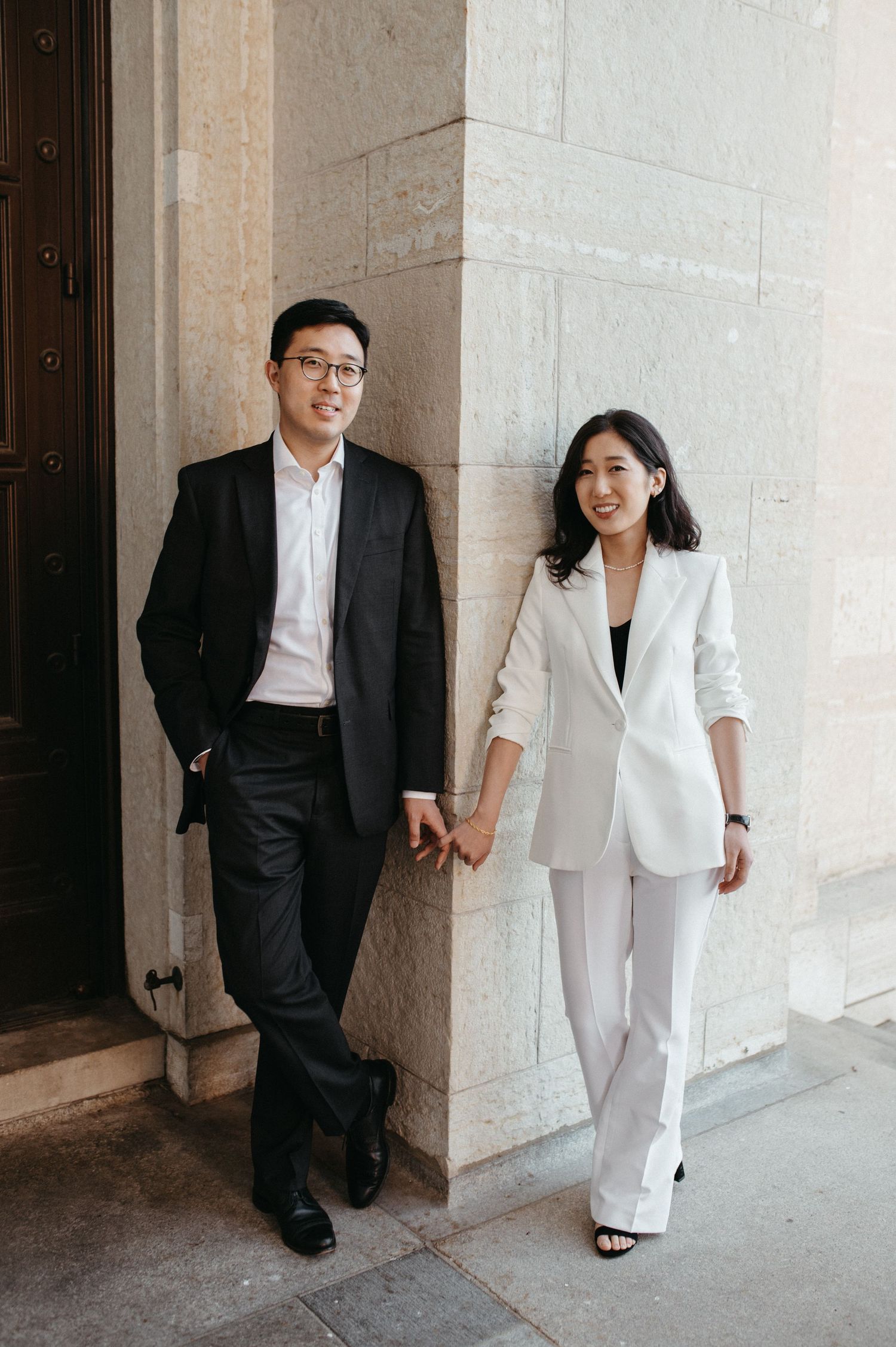 Contemporary couple in contrasting suits posing against light stone architecture.