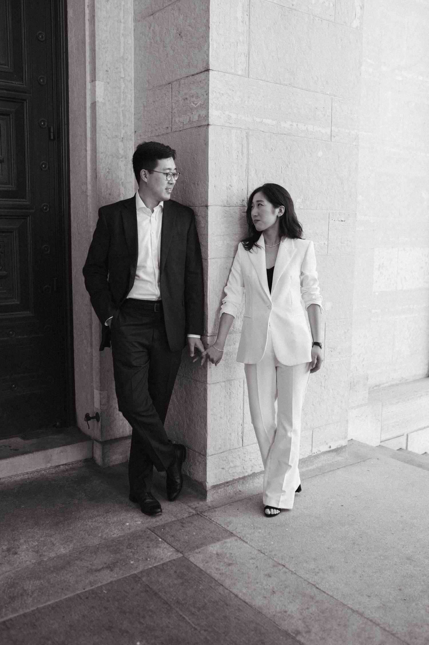 Black and white photo of a couple in business attire leaning against a stone wall.
