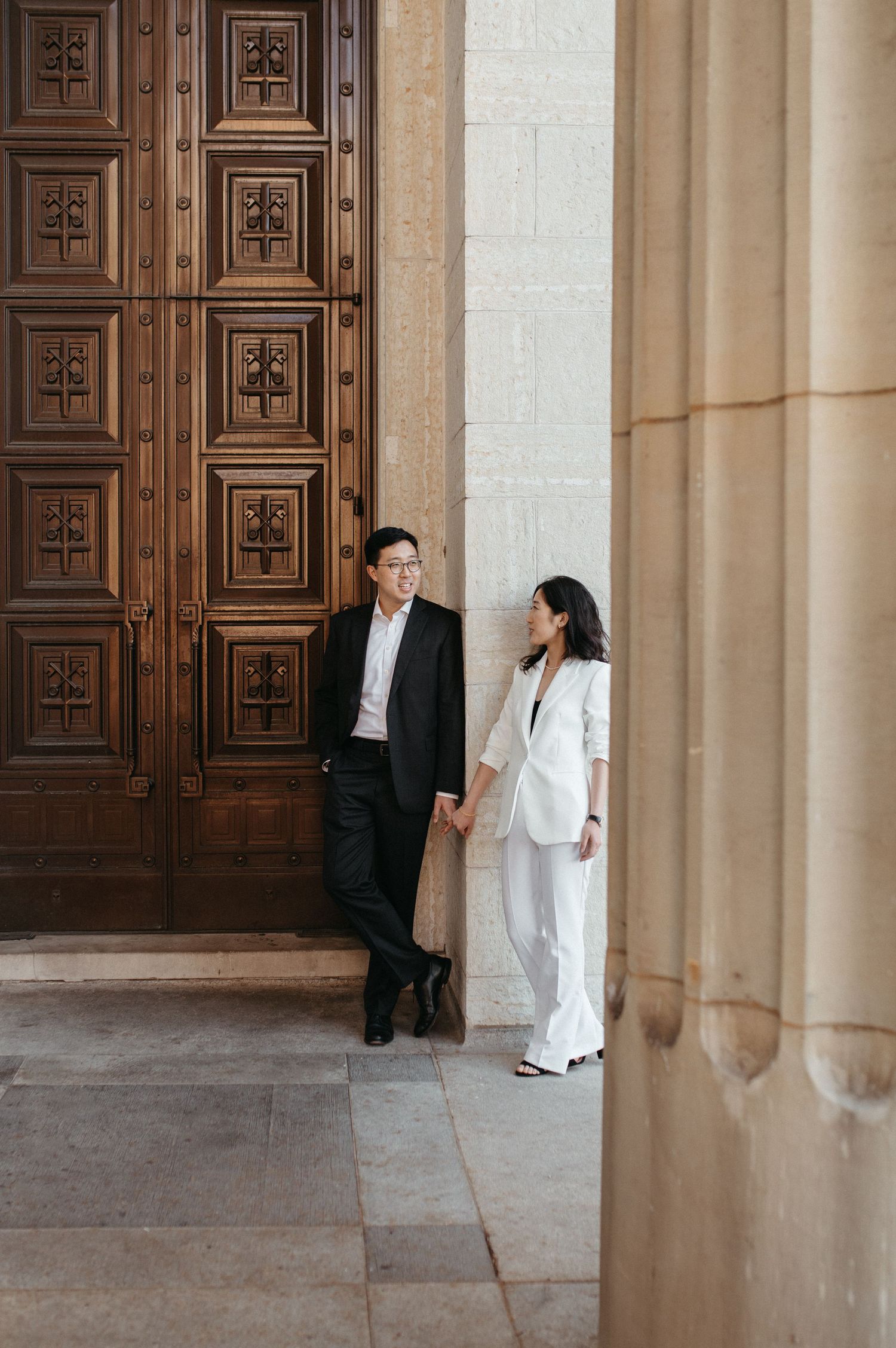 Elegant couple posing by ornate wooden doors in a classical architectural setting.