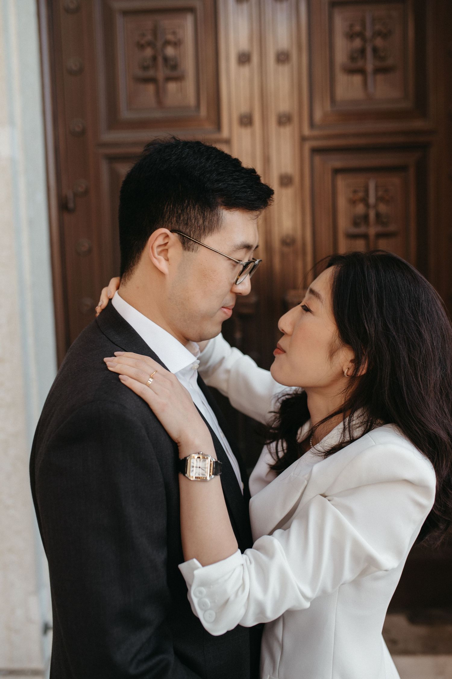 An intimate couple in formal attire embrace in front of ornate wooden doors.